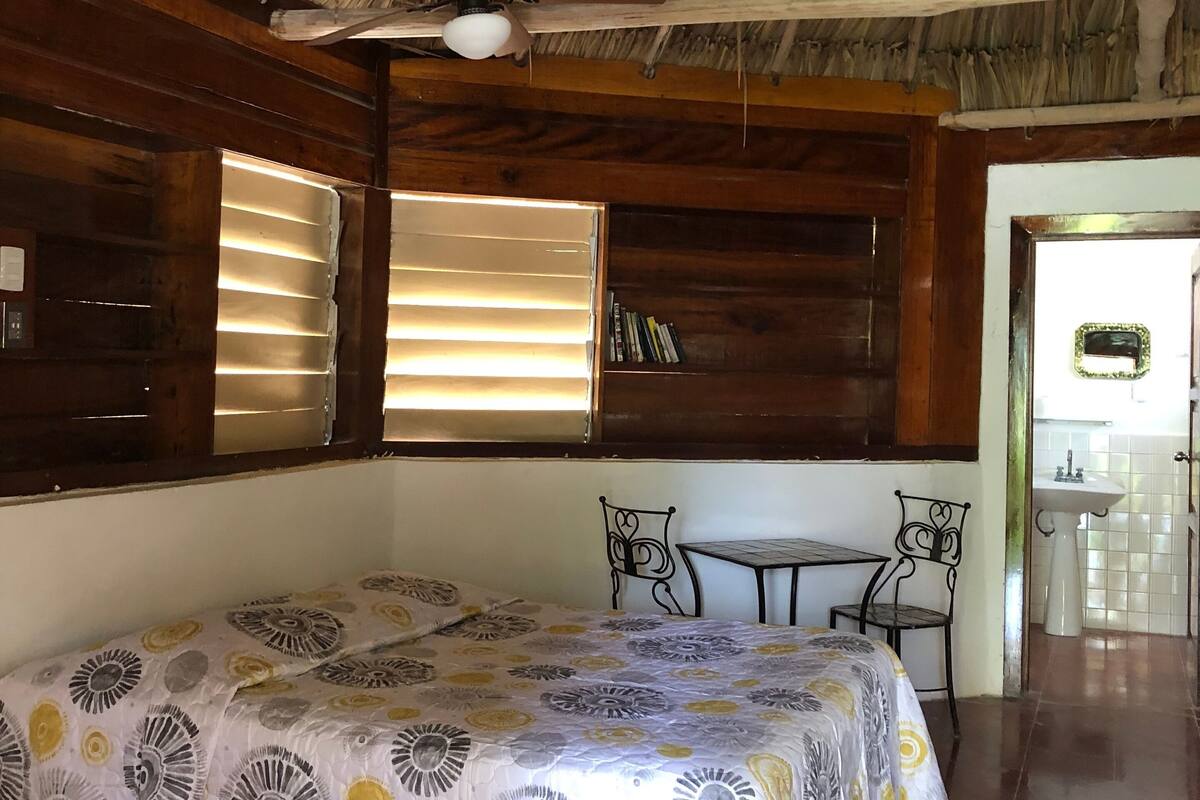 Ein Zimmer mit einem Bett, einem Tisch und einem Stuhl. Es gibt einen Deckenventilator und ein Fenster mit Jalousien.