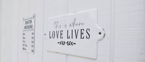 A white wall with two signs: one labeled 'BATH HOUSE' listing prices for various services, and another stating 'this is where LOVE LIVES' with decorative leaf designs.
