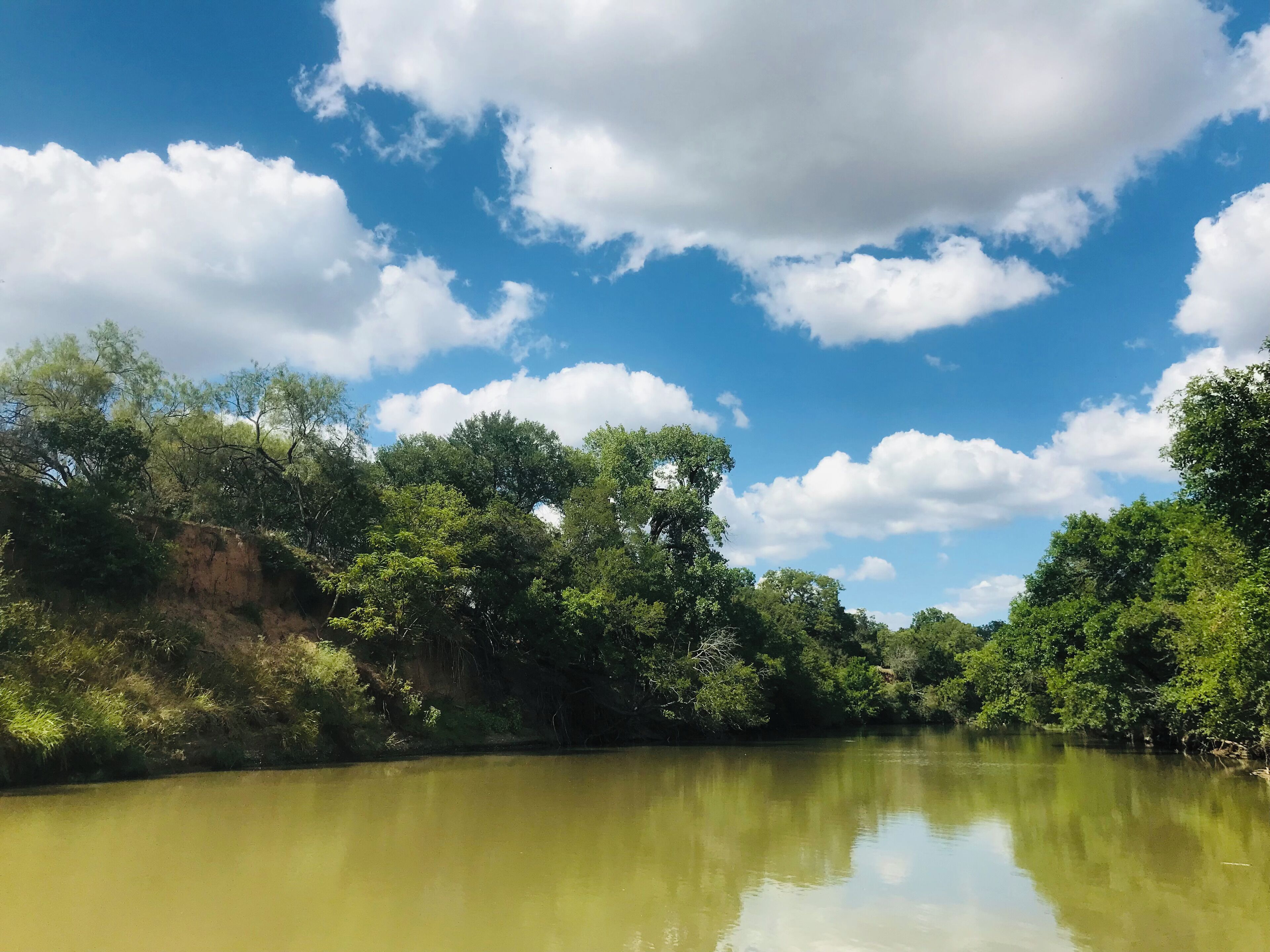 Un río con abundante vegetación verde a ambos lados, cielo azul claro con nubes dispersas y una superficie acuática tranquila que refleja el cielo.