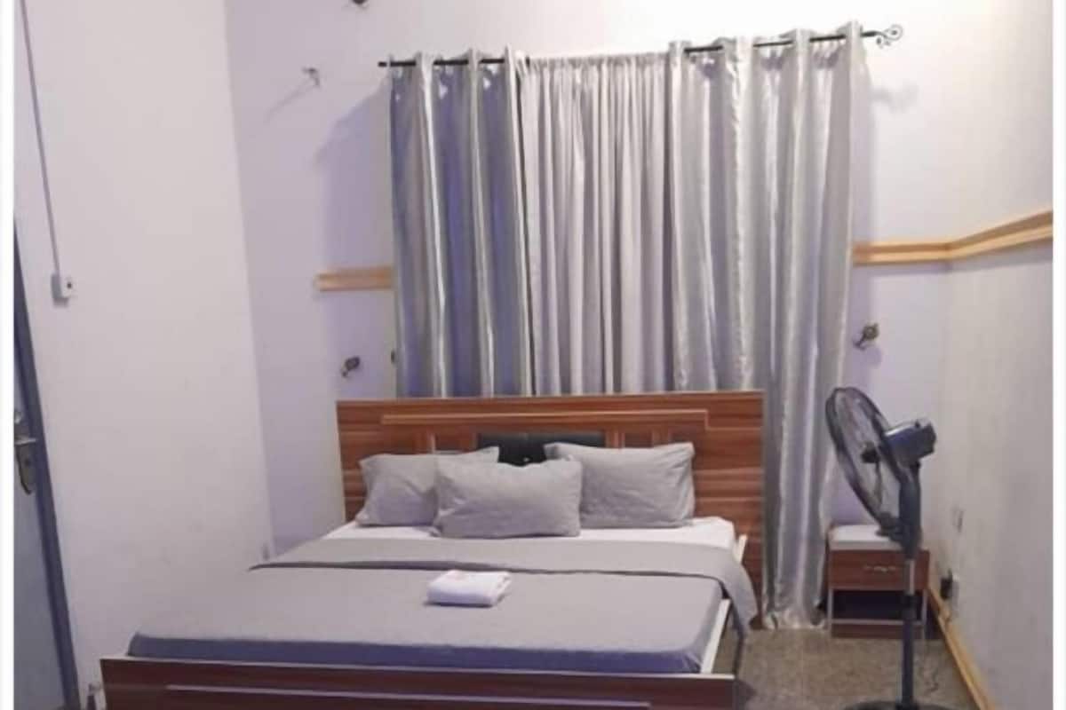 Een slaapkamer met een bed, nachtkastje, ventilator en gordijn.