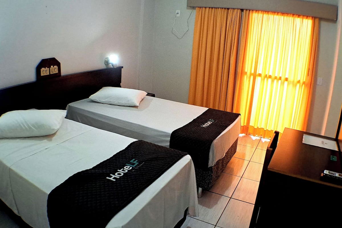 Habitación de hotel con dos camas, una mesita de noche, una lámpara y una ventana con cortinas.