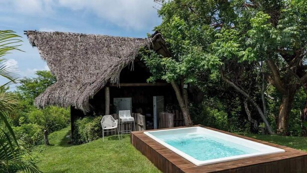 Une cabane au toit de chaume, dotée d’une terrasse en bois et d’un jacuzzi, nichée dans un jardin luxuriant.
