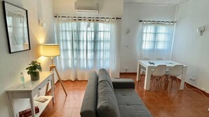A living room with a grey sofa, a small white table with a lamp, a dining area with a white table and chairs, and a ceiling with a textured pattern. - Grupo Morgado (Tavira)