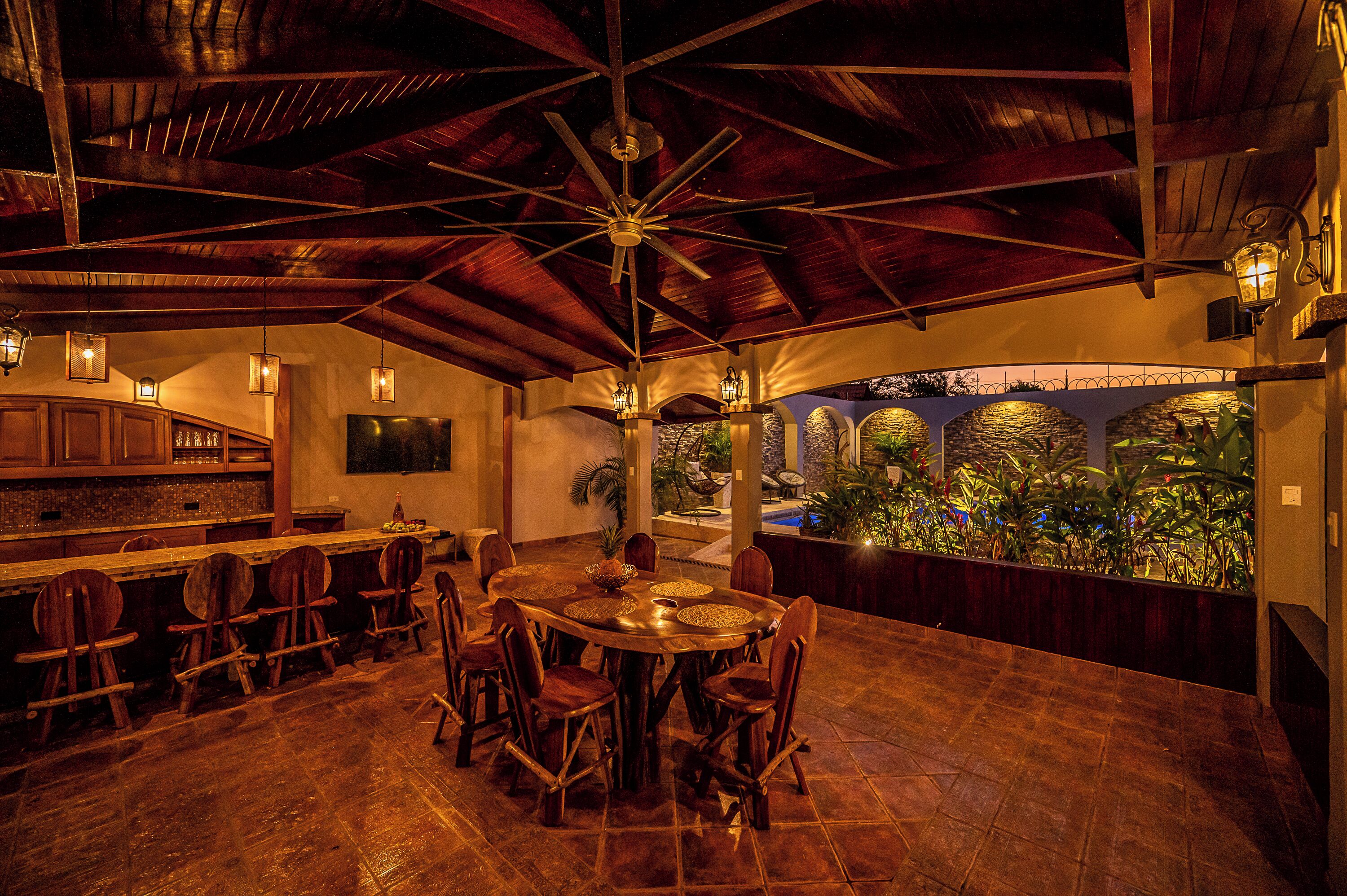 Ein Restaurant mit hohen Decken, Holzbalken und einem zentralen Ventilator. Es gibt Holzstühle und einen runden Tisch mit einer gewebten Tischmatte. Der Boden ist mit Fliesen ausgelegt, und es gibt einen Barbereich mit Holzsärchen und einem Fernseher.