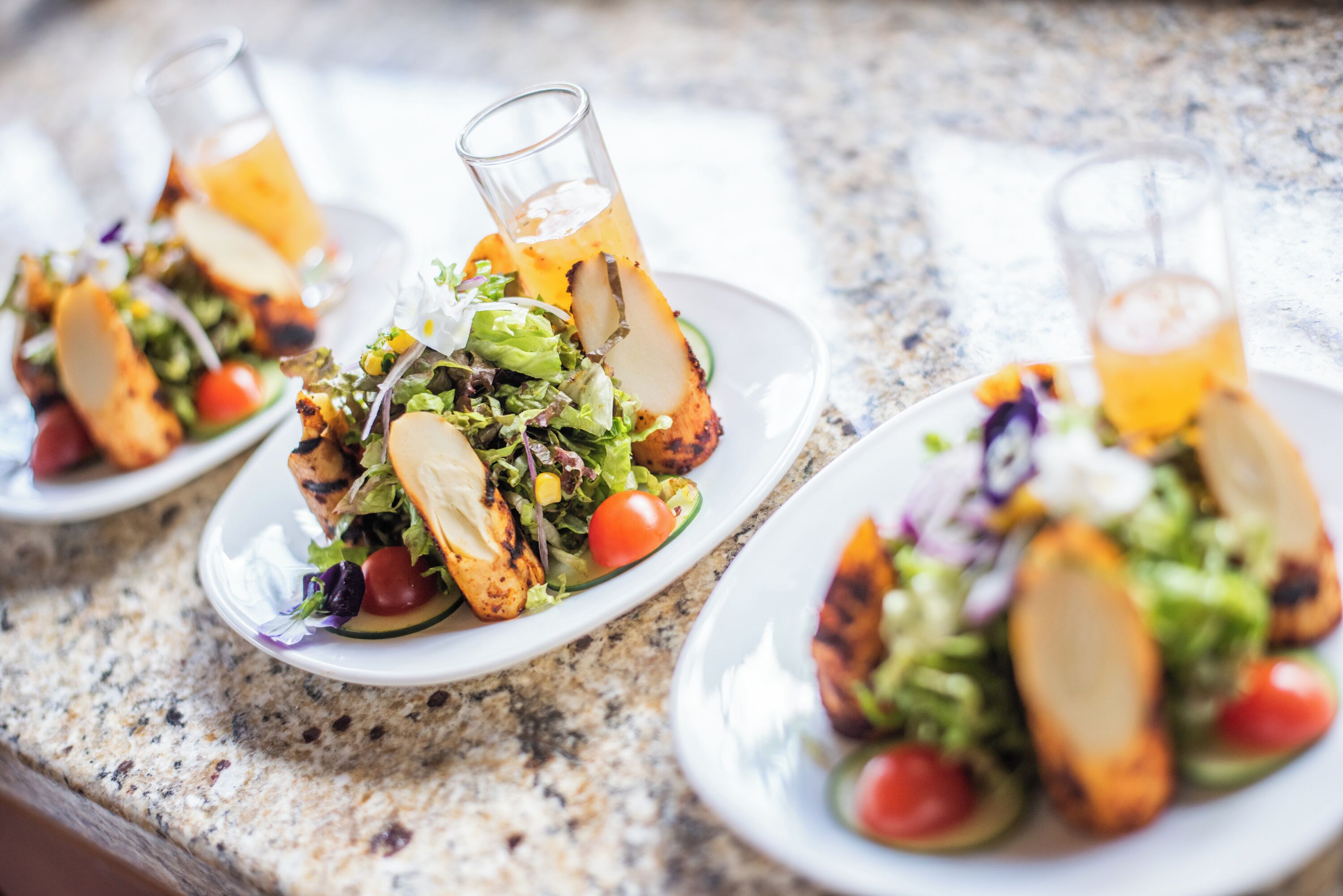 Three plates of salad with grilled chicken and a glass of orange juice on a granite countertop.