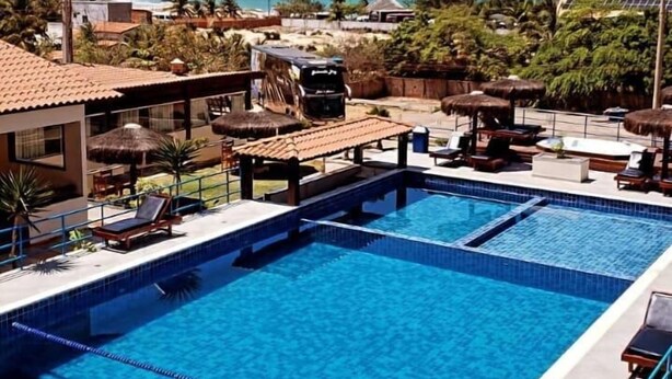 A swimming pool with lounge chairs and umbrellas, surrounded by palm trees and a view of the ocean. - Hotel Pousada Canoa Quebrada (Aracati)