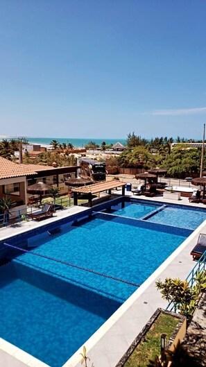 A swimming pool with lounge chairs and umbrellas, surrounded by palm trees and a view of the ocean. - Hotel Pousada Canoa Quebrada (Aracati)