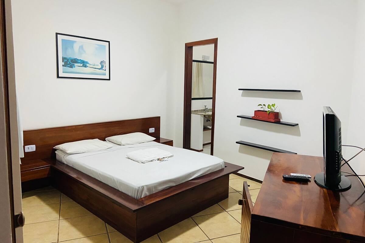 A hotel room with a wooden bed, a small desk with a TV, a shelf with a plant, and a framed picture on the wall.