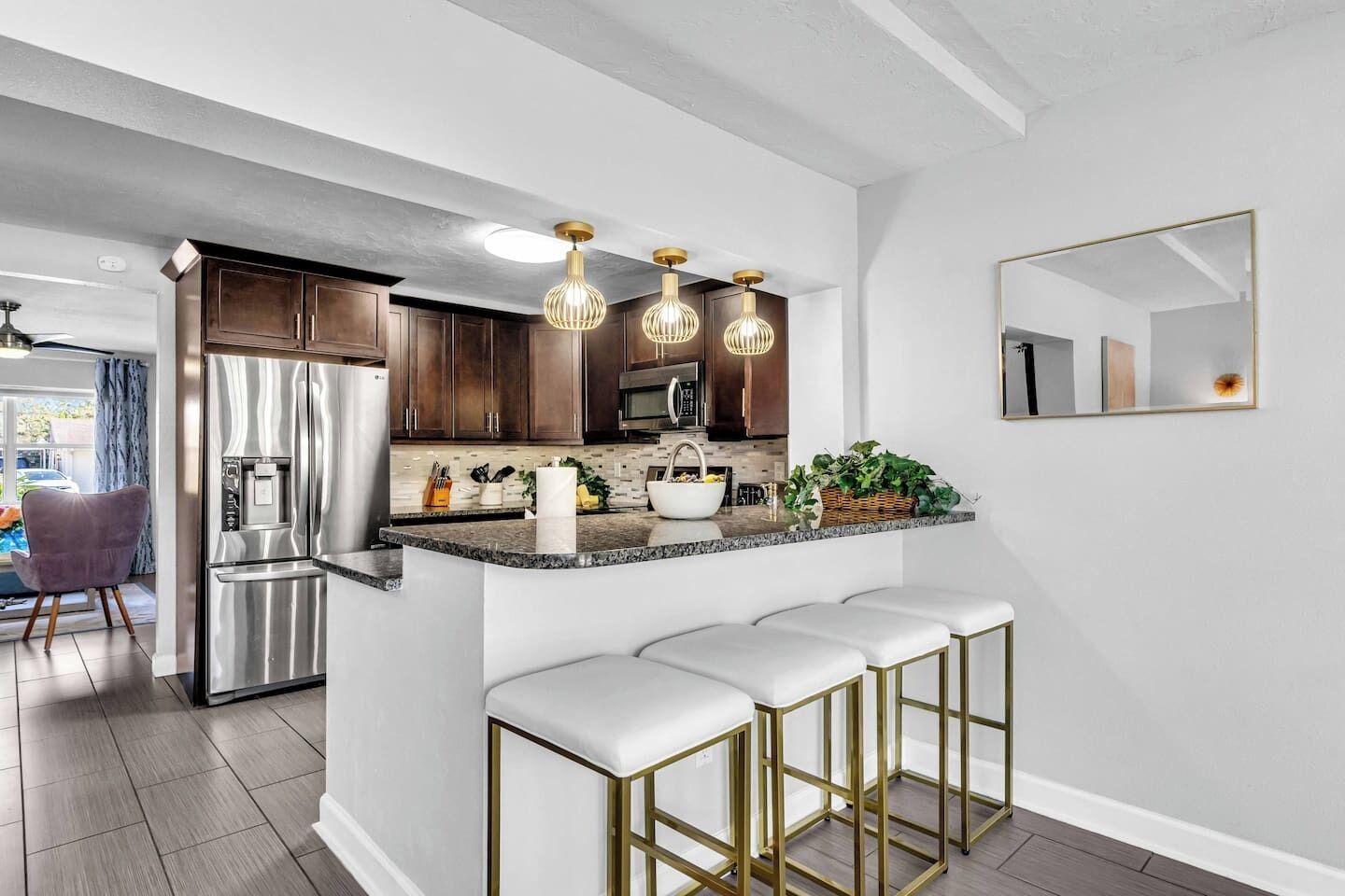 A modern kitchen with dark wood cabinets, stainless steel appliances, and a marble countertop. There are three bar stools with gold frames and white cushions.
