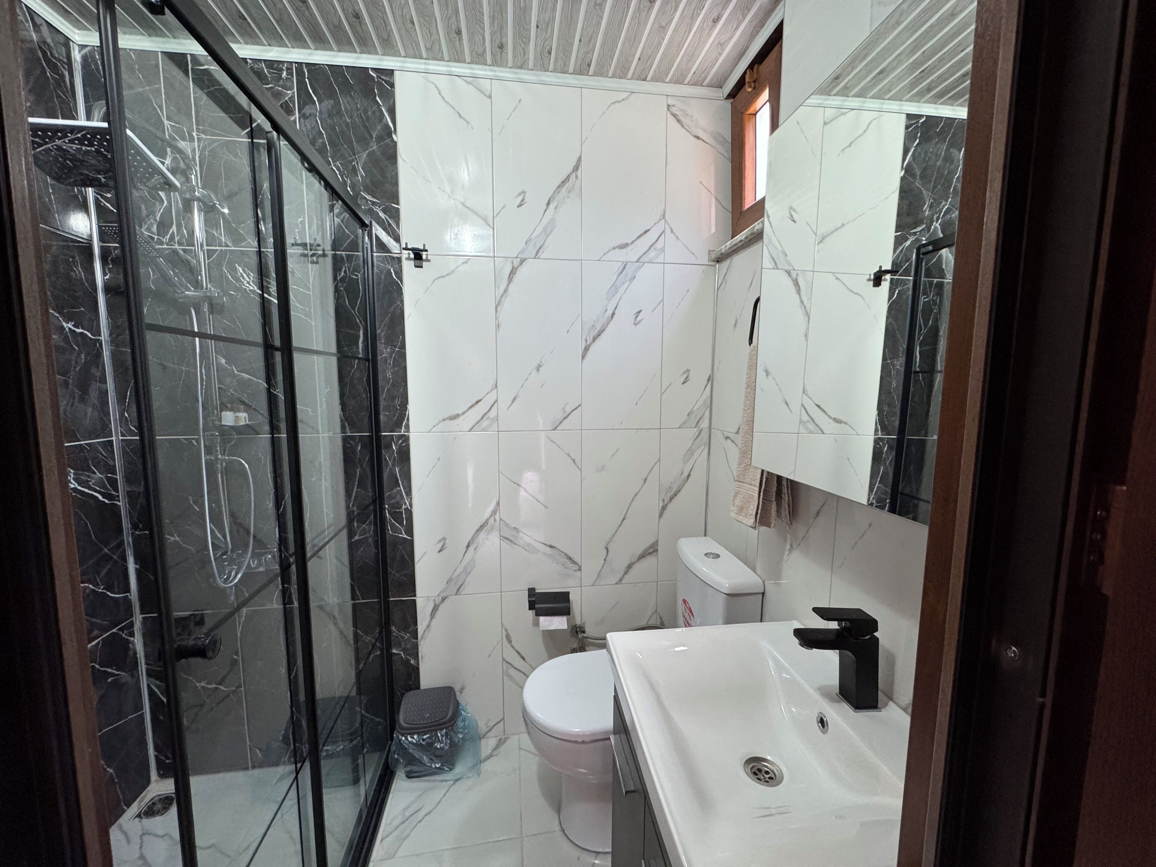 Une salle de bain dotée d’une douche à parois de verre, de carreaux de marbre blanc, de toilettes blanches et d’un lavabo avec un robinet noir.