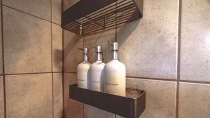 Une salle de bain avec une étagère sur laquelle sont posées trois bouteilles de produits de toilette : gel douche, shampoing et après-shampoing.
