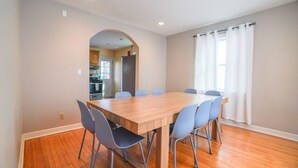 Un coin repas avec une table en bois et des chaises bleues, une cuisine visible à travers une arche, et une fenêtre ornée de rideaux blancs.