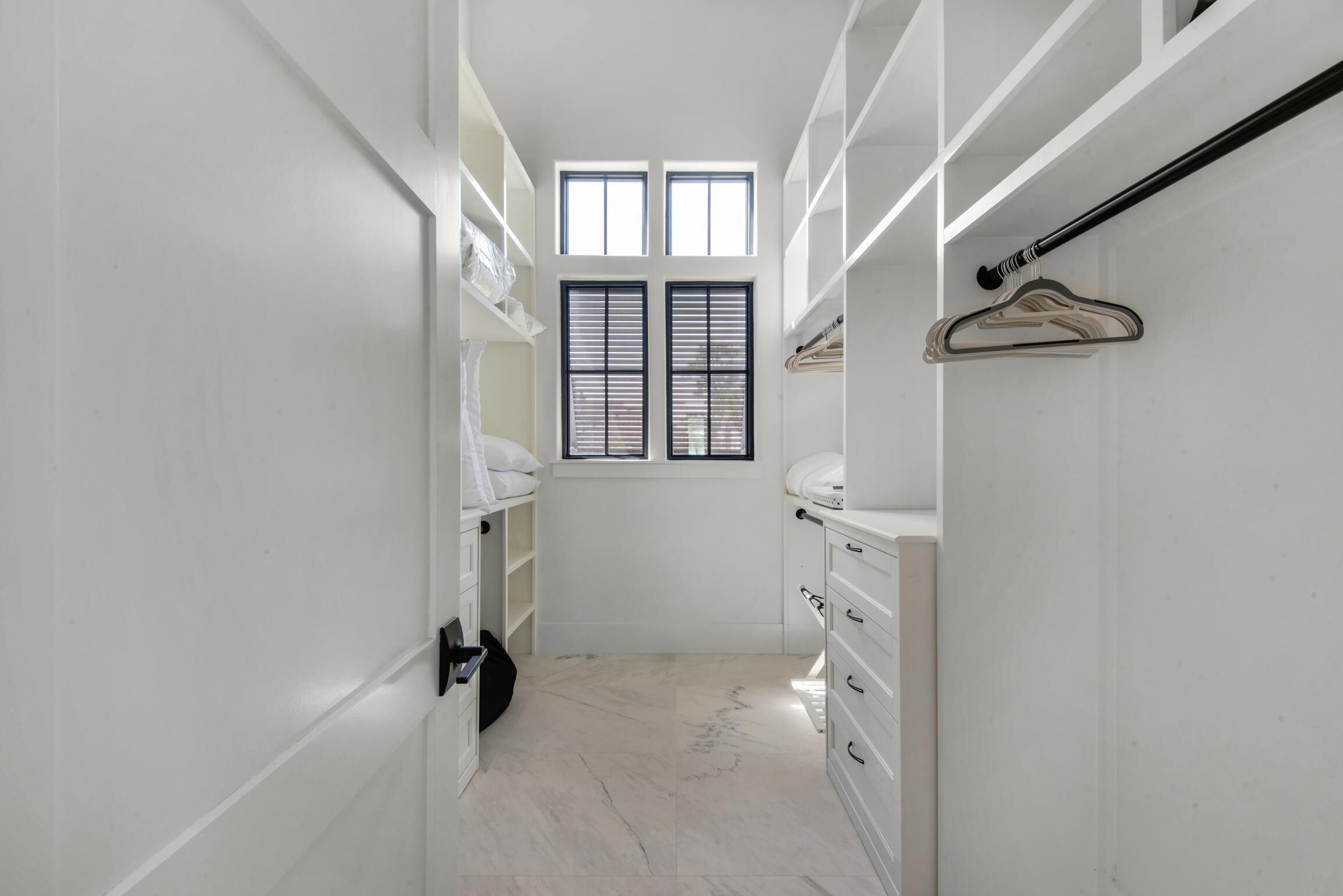 A narrow walk-in closet with white cabinetry, a hanging rack, and a window with blinds.