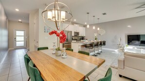 Un grand espace à manger avec une table en bois et des chaises vertes, une cuisine moderne en arrière-plan et un coin salon avec un canapé et une télévision.