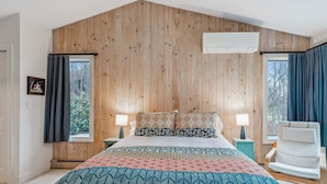 Een slaapkamer met een houten wand, een bed met beddengoed met een patroon, twee nachtkastjes met lampen en een stoel bij een raam.