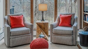 Een woonkamer met twee fauteuils, een klein bijzettafeltje, een lamp en een groot raam met uitzicht op bomen.