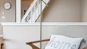 Une pièce avec une chaise en bois, une lampe, un escalier et une horloge murale.