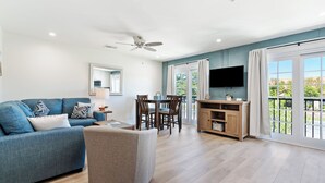 Un salon moderne avec un canapé bleu, un fauteuil gris, une table à manger avec des chaises, un ventilateur de plafond, un téléviseur fixé au mur et de grandes fenêtres avec des rideaux.