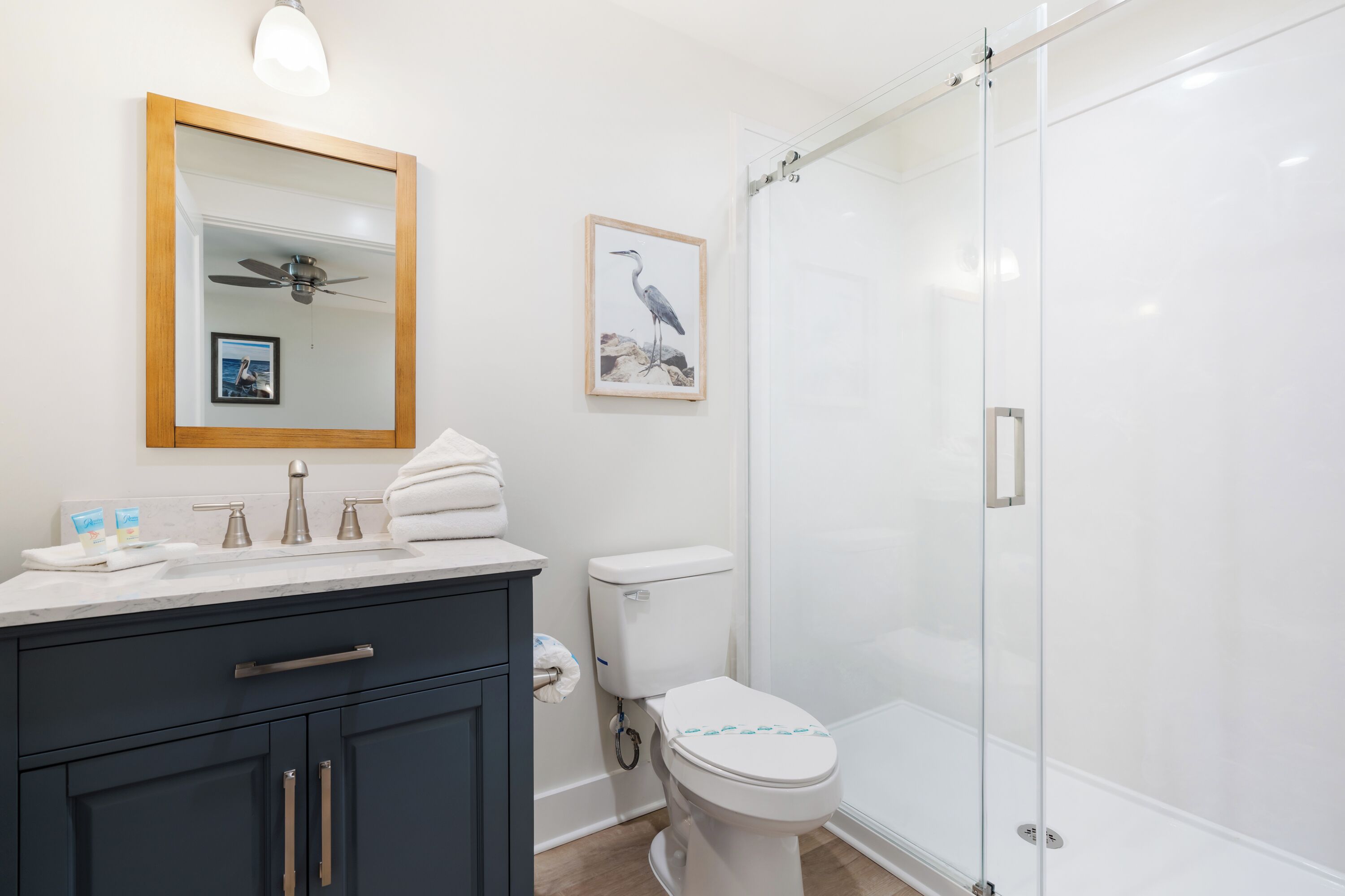 A bathroom with a dark vanity, a white toilet, a glass shower enclosure, and a ceiling fan.