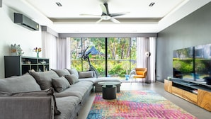 A modern living room with a grey sofa, a colorful rug, a TV, and a balcony with a view of greenery. - Laguna Park 4 Bedrooms Villa by NLA (Choeng Thale)