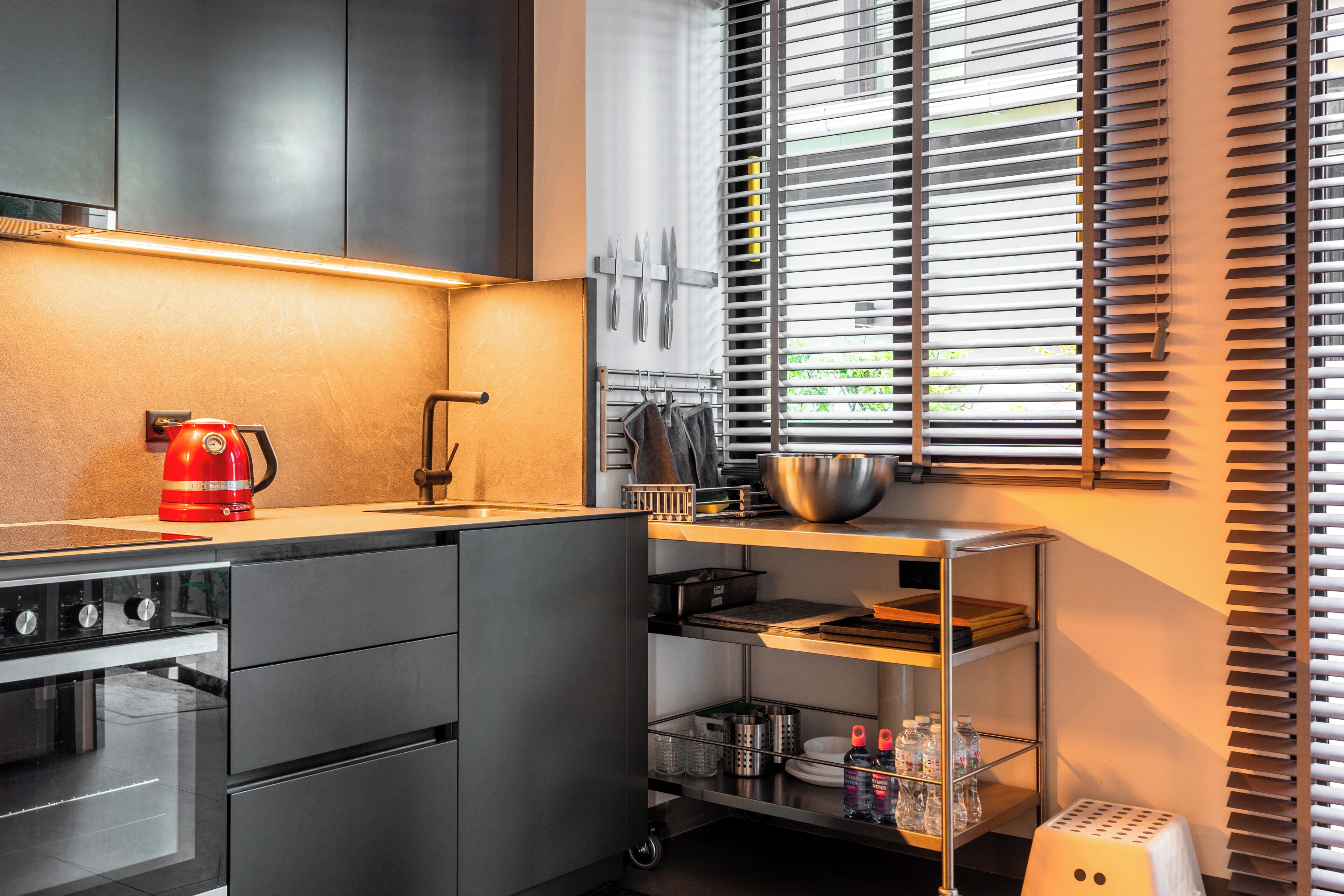 A modern kitchen with a stove, oven, and a red kettle. There is a metal shelf with kitchen utensils and a window with blinds.