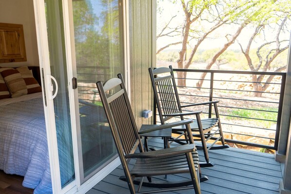 Una habitación con cama, puerta corrediza de vidrio que da a un espacio exterior con sillas y vistas a árboles.
