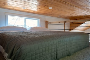 Un dormitorio con techo de madera, una cama con colcha verde y una ventana con vista a árboles.
