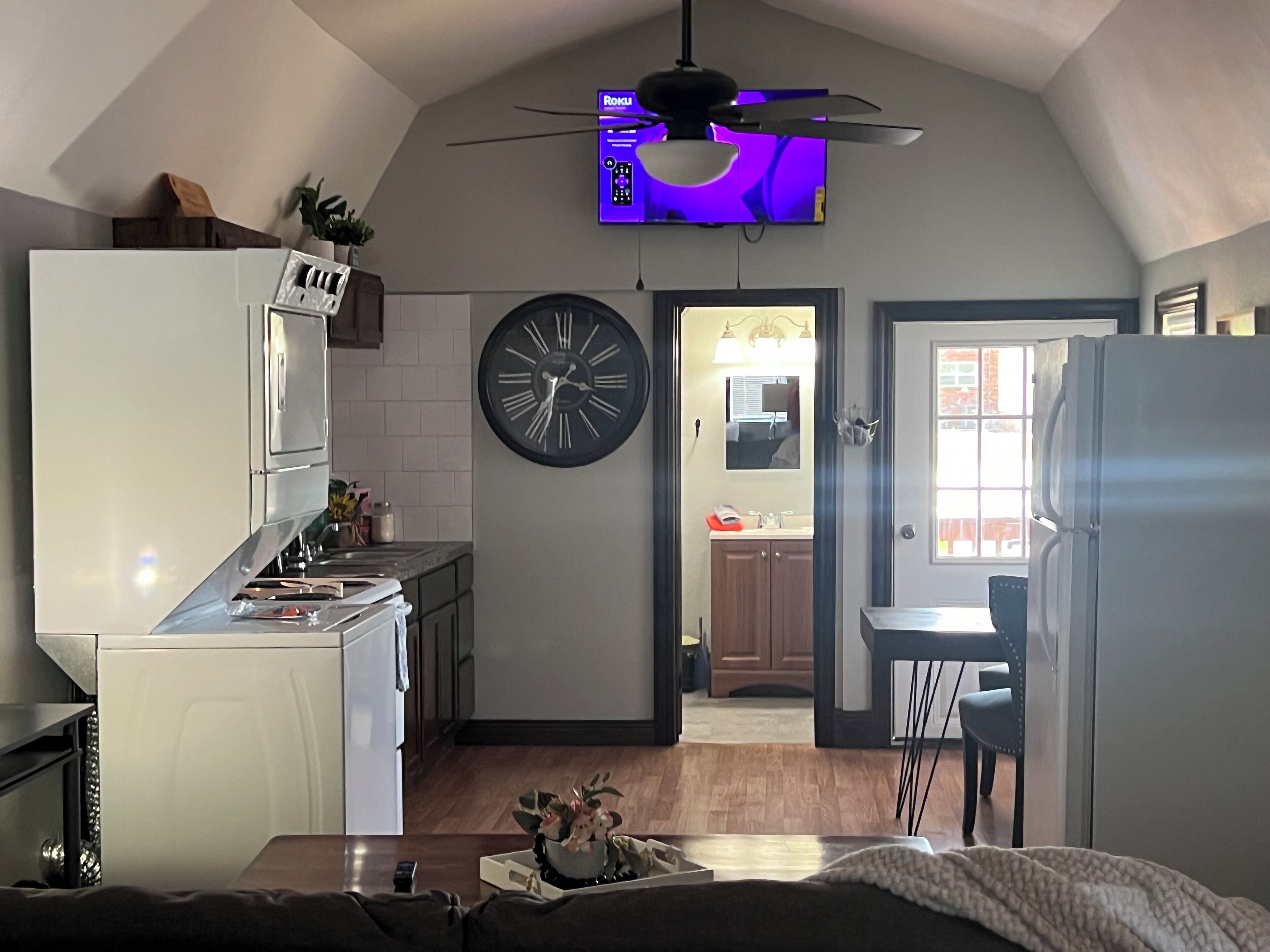 Une cuisine de taille réduite, équipée d’une horloge, d’un ventilateur de plafond et d’un téléviseur fixé au mur.