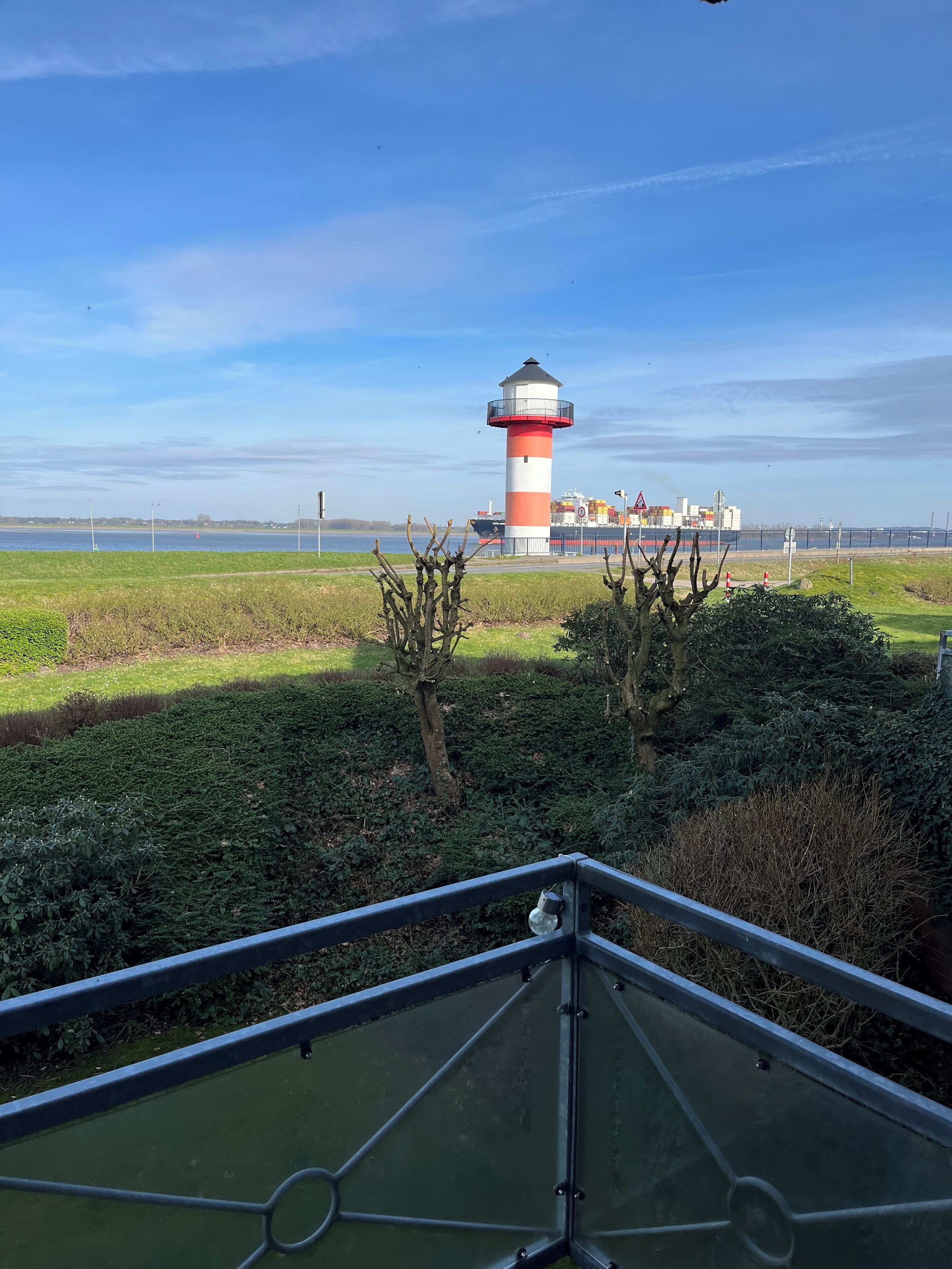 Une vue d’un phare depuis un balcon doté d’une rambarde en métal, avec un champ herbeux et une étendue d’eau en arrière-plan.