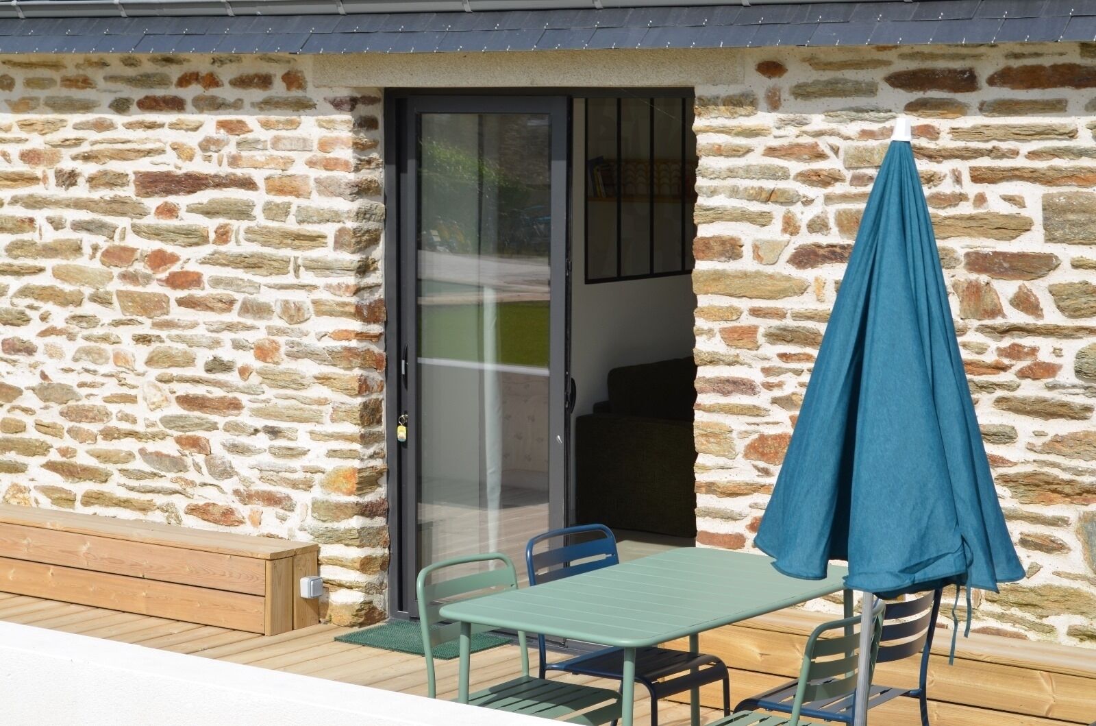 A stone building with a wooden deck, a table with chairs, and a blue umbrella.