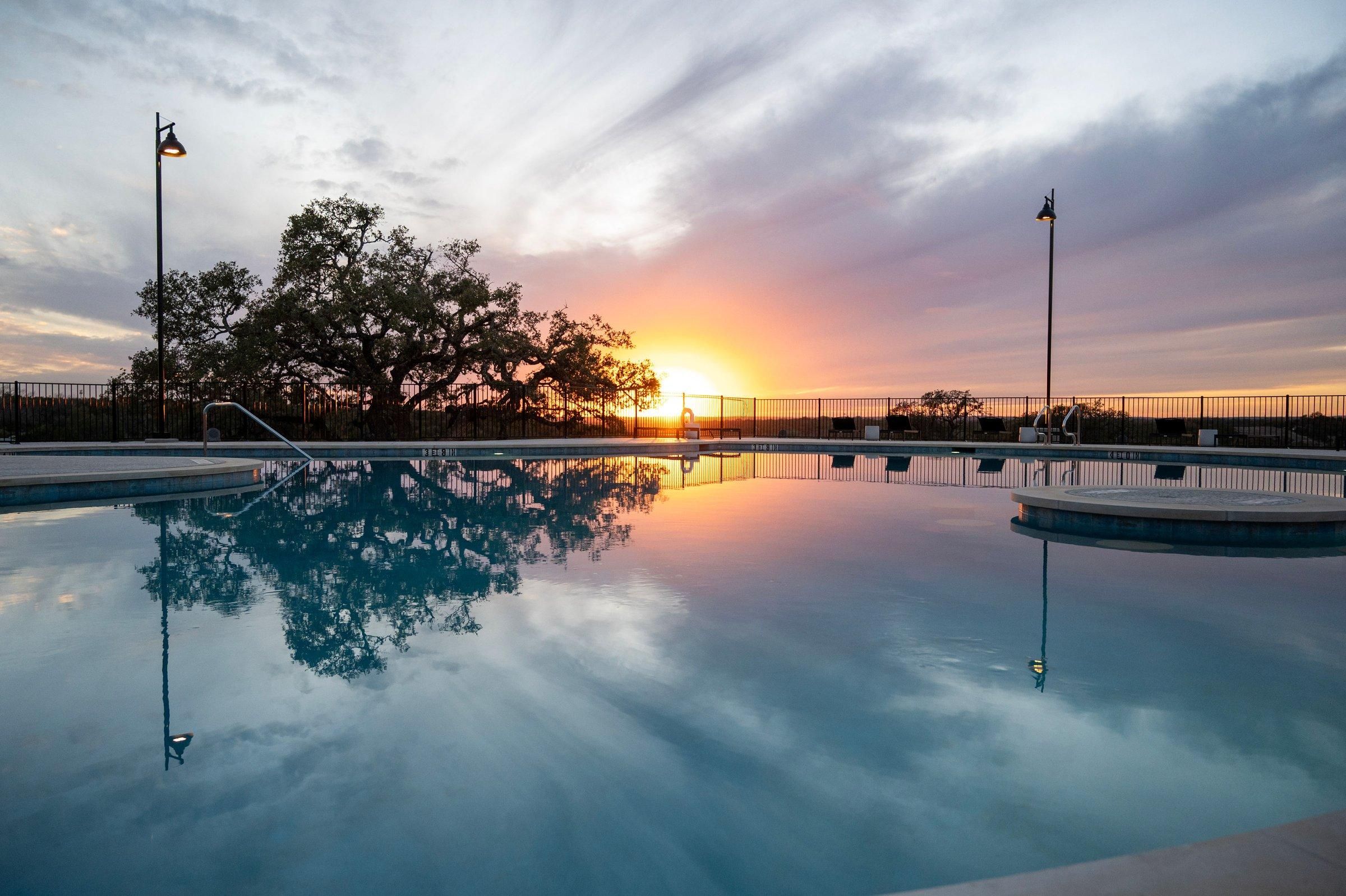 A swimming pool with a sunset view, surrounded by a fence, and two street lamps.