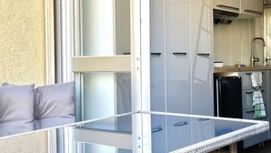 Un balcon avec une table en verre, un canapé avec des coussins et un coin cuisine équipé de placards et d’un réfrigérateur.