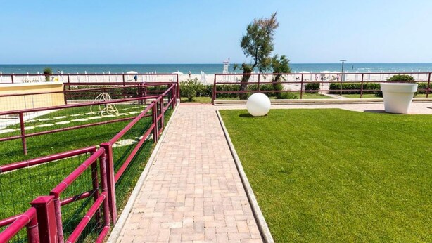 Uma passarela pavimentada com guarda-corrimão vermelho, área gramada e vista para o mar.