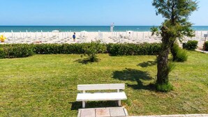 Uma área gramada com um banco branco, uma árvore e uma praia com guarda-sóis ao fundo.