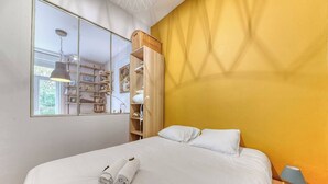 Un dormitorio con una cama, almohadas blancas, una mesita de noche con lámpara, un espejo y un estante con libros y una cesta.