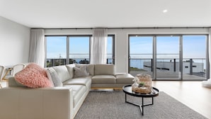 A modern living room with a sectional sofa, a coffee table, and large windows offering a view of a waterfront and buildings. - Casa La Pose San Remo gateway to Phillip Island (San Remo)