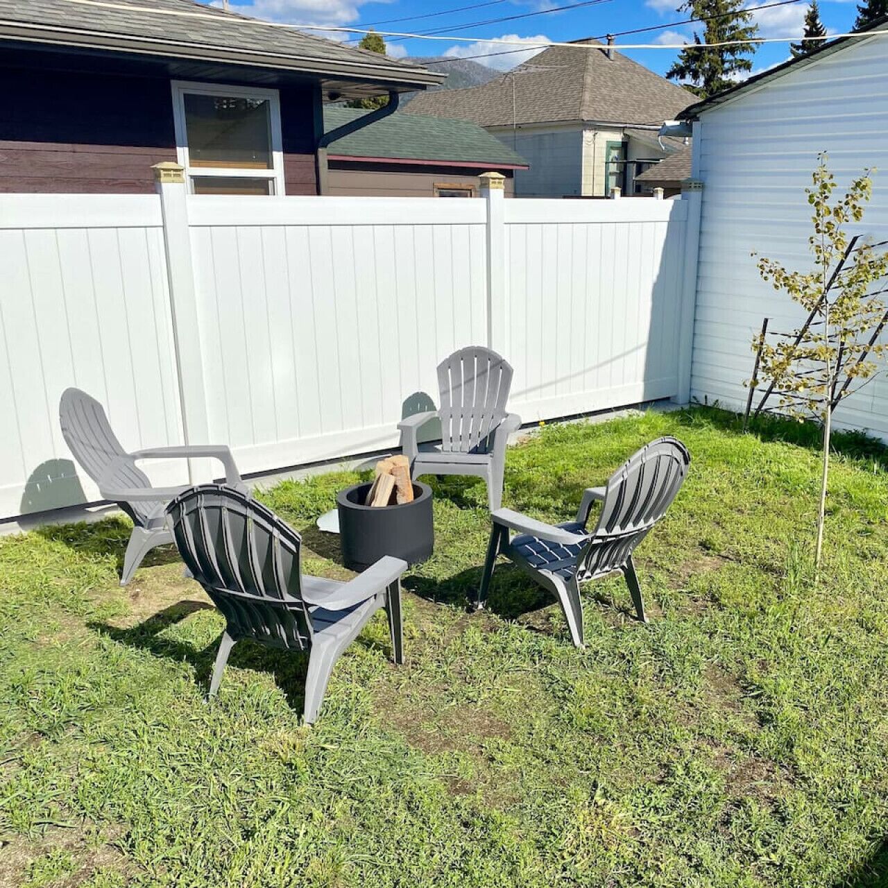 Un jardin avec une clôture blanche, trois fauteuils Adirondack, un foyer noir et un petit arbre.