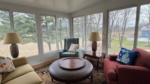 A sunroom with large windows, a round coffee table, a red sofa, a blue armchair, and two lamps.