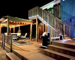 Una terraza de madera con zona de estar cubierta, una escalera y luces colgantes.