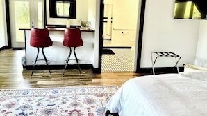 A modern hotel room with a large bed, a sitting area with two red chairs, a small desk, and a bathroom visible through an open door.