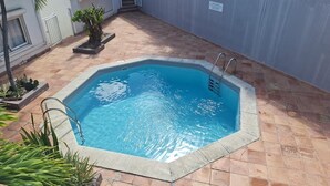 A rectangular swimming pool with a ladder on one side, surrounded by a tiled patio with a few potted plants.