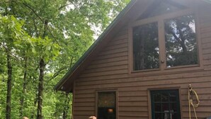 A two-story wooden cabin with a deck, surrounded by trees. - Quaint Cabin in the Trees!
 (Counce)