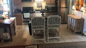 A kitchen with wooden cabinets, a stainless steel refrigerator, a microwave, and a dining area with two chairs. - Quaint Cabin in the Trees!
 (Counce)