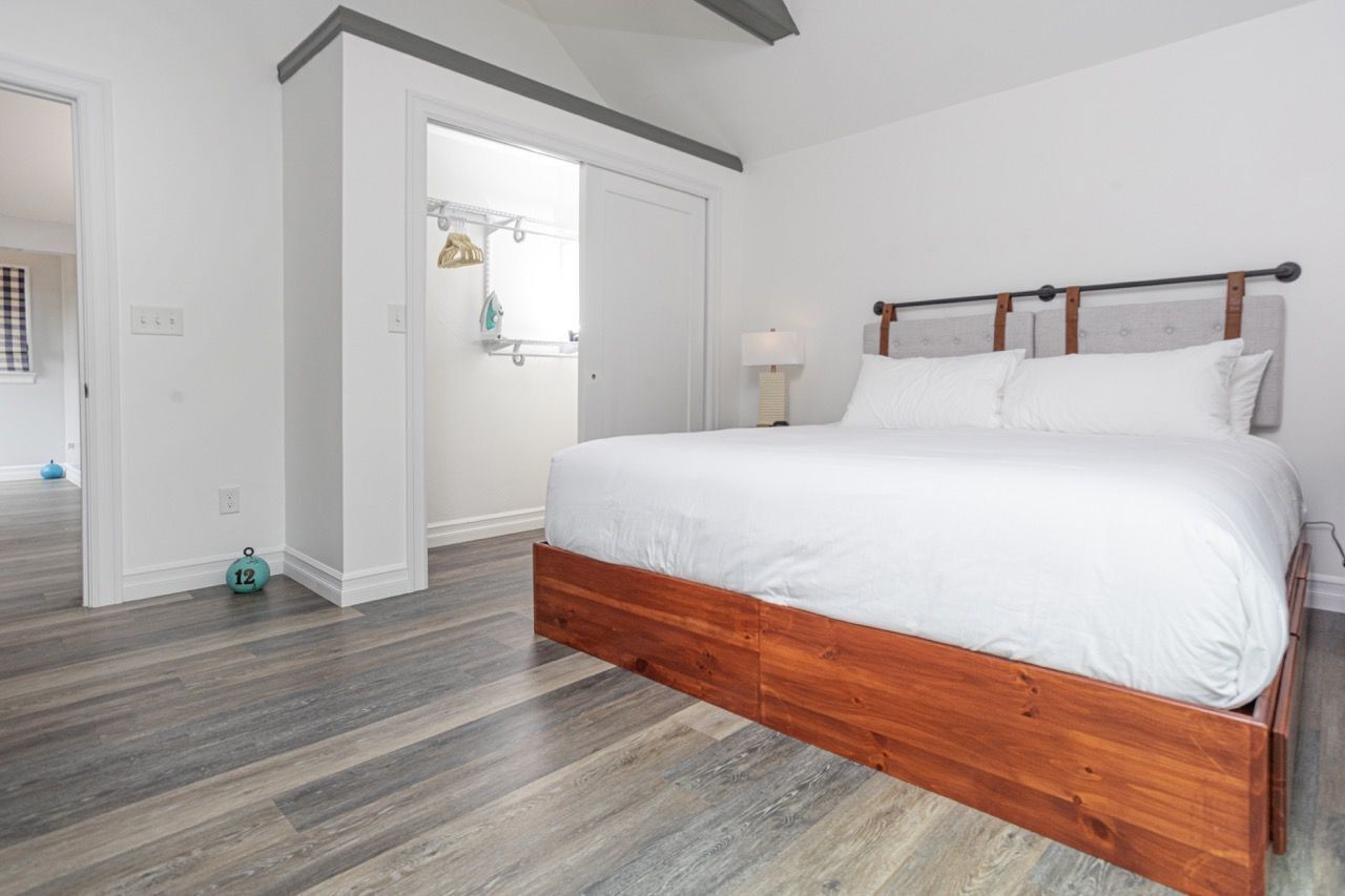 A bedroom with a wooden bed, white bedding, a nightstand, a lamp, and a door leading to another room.