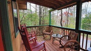 Terraza de madera con mecedoras y una mesa metálica, con vista a un área boscosa.