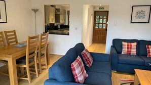 A modern living room with a blue sofa, wooden dining table, and chairs. There is a kitchen visible through an open door. - Selwood Cottage - A peaceful, rural retreat on the outskirts of Frome, close to Longleat Safari Park (Beckington)