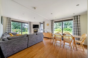 A modern living room with a grey sectional sofa, wooden dining table with chairs, a flat-screen TV mounted on the wall, and large windows offering a view of the garden. - Farleigh Lodge - A peaceful,  rural retreat on the outskirts of Frome, near Longleat Safari Park and (Beckington)