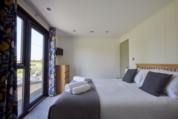 A bedroom with a large bed, a wooden headboard, a window with curtains, a bedside table, and a flat-screen TV mounted on the wall. - Farleigh Lodge - A peaceful,  rural retreat on the outskirts of Frome, near Longleat Safari Park and (Beckington)