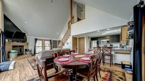 A spacious living area with a dining table set for four, a kitchen in the background, and a staircase leading to an upper level.