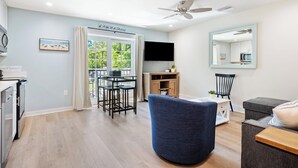 A modern living room with a dining area, a ceiling fan, a flat-screen TV, and a large window with curtains.